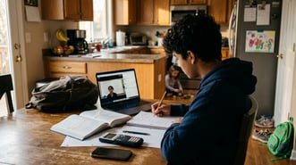A 9th grade boy working through an algebra problem at a kitchen table with a notebook and pencil.